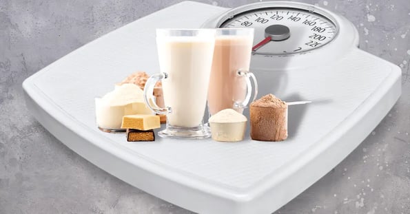 A weight scale with protein shakes, powder, and bars stacked on top of it on a gray background.